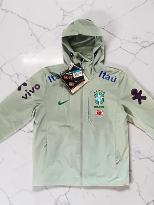 Ljusgrön Brasilien vindjacka Nike - Ljusgrön vindjacka från Nike med Brasilien landslagslogga på bröstet, flera sponsortryck i blått och lila samt grönt Nike-märke. Jackan har huva, dragkedja och två sidofickor. Materialet är GORE-TEX för extra skydd mot vind och regn.