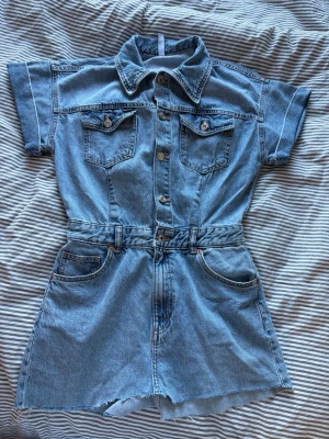 Kort jeansklänning med knappar - Säljer en kort jeansklänning i blå denim med korta ärmar, krage och knappar framtill. Klänningen har två bröstfickor med lock och knapp samt två sidofickor. Perfekt för en avslappnad och cool stil. DETTA HAR FRÅN BÖRJAN VARIT EN JENS JUMPSUITE MEN ÄR AVKLIPPT TILL EN KORT KLÄNNING☺️