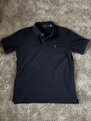 Mörkblå polotopp från Polo Ralph Lauren - Klassisk mörkblå polotopp från Polo Ralph Lauren med två knappar och krage. Toppens material är bomull och den har ett broderat rött logomärke på bröstet. Perfekt för en stilren look och enkel att matcha med jeans eller shorts.