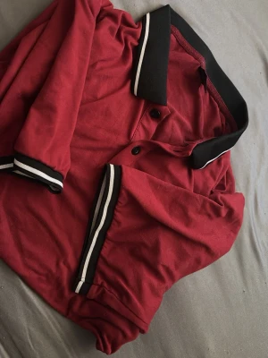 Röd pikétröja med svarta detaljer - Säljer en röd pikétröja med svart krage och svarta muddar med vita ränder. Tröjan har knappar framtill och är tillverkad i mjuk bomull. Perfekt till en avslappnad och stilren look.