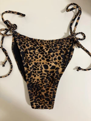 Leopardmönstrad bikiniunderdel med knyt - Snygg bikiniunderdel med leopardmönster och knyt i sidorna. Färgerna går i brunt och svart, vilket ger en trendig och djurisk look. Perfekt för strandhäng eller poolparty. Materialet känns mjukt och följsamt mot huden.