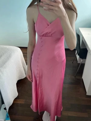 Rosa satinklänning med knappar - Säljer en rosa satinklänning med smala axelband och omlott-ringning. Klänningen har dekorativa knappar längs sidan och en lätt utställd midilång kjol. Materialet är glansigt och följsamt, vilket ger en elegant look.