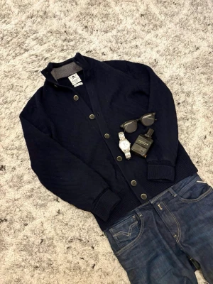 Mörkblå cardigan från Hackett - Snygg mörkblå cardigan från Hackett med klassisk knappstängning och ribbade muddar. Size S och passar upp till längden 175.