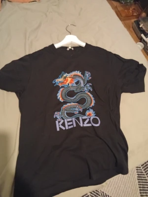 Svart Kenzo t-shirt med drakmotiv - Cool svart t-shirt från Kenzo med ett stort färgstarkt drakmotiv på framsidan och Kenzo-loggan under. T-shirten har korta ärmar och är gjord i mjuk bomull, perfekt för dig som vill sticka ut med en unik design.