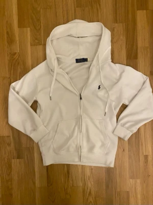 Vit hoodie från Polo Ralph Lauren - Vit hoodie med dragkedja från Polo Ralph Lauren. Klassisk design med två stora fickor framtill, justerbar huva och broderad svart logotyp på bröstet. Mjuk bomullskvalitet som är riktigt skön att ha på sig.