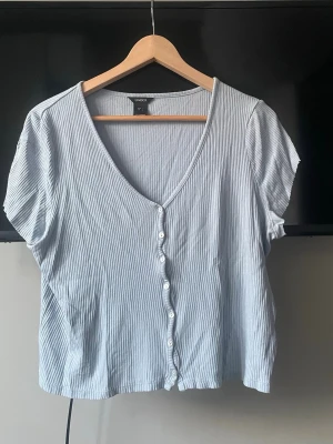 Randig kortärmad topp  - Säljer en ljusblå och vit randig topp från Lindex med v-ringning och knappar framtill. Toppen är kortärmad och har en ribbad struktur som ger en snygg och avslappnad look. Perfekt för dig som gillar stilrena och bekväma plagg. Ärmarna är avklippta men annars så är tröjan i ett väldigt bra skick. Storlek L men passar s/m också❣️