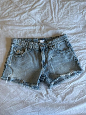 Ljusa denimshorts med broderade fickor - Säljer ett par ljusa denimshorts med råa kanter och slitningar. Shortsen har broderade bakfickor med snygga detaljer och klassisk femficksdesign. Perfekta för sommarens alla äventyr, och de har en normal passform och midwaist.