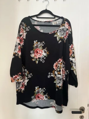 Svart blommig långärmad tröja  - En svart blus med stora blommiga mönster i rosa, grått och vitt. Blusen har rund halsringning och trekvartsärmar.