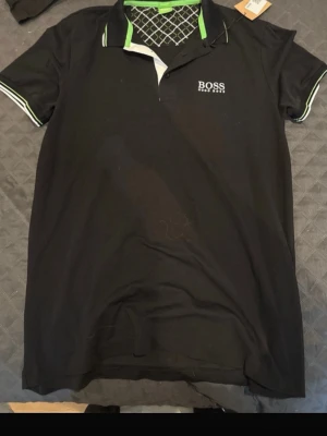 Svart pikétröja från BOSS - Snygg svart pikétröja från BOSS med vit och grön detalj på kragen och ärmsluten. Tröjan har klassisk krage, korta ärmar och BOSS-logga broderad på bröstet och i nacken. Tillverkad i mjukt bomullsmaterial för en bekväm känsla.