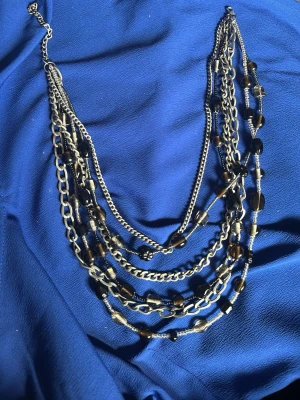 Flerradigt halsband med svarta pärlor - Snyggt flerradigt halsband med silverfärgade kedjor och svarta pärlor. Kedjorna har olika länkar och pärlorna är både runda och avlånga, vilket ger ett coolt och trendigt uttryck. Perfekt för att lyfta din outfit med en edgy touch.