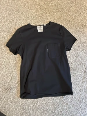Svart t-shirt med bröstficka - En stilren svart t-shirt från Cheap Monday med en enkel bröstficka på vänster sida. T-shirten har rund hals och korta ärmar, perfekt för en avslappnad look. Materialet känns mjukt och skönt mot huden. Stl 34 är jag ej säker på att det är men annars är det stl S