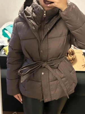 Brun pufferjacka med bälte och huva - Säljer en brun pufferjacka med bälte i midjan och stor huva. Jackan har dragkedja och knappar framtill, quiltad design och är tillverkad i ett glansigt material som håller dig varm under kalla dagar. Perfekt för dig som vill ha en trendig och bekväm vinterjacka.