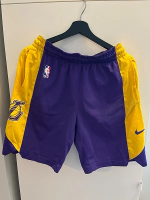 Lakers basketshorts Nike Dri-FIT - Säljer ett par Lakers basketshorts från Nike med Dri-FIT material. Shortsens färg är lila och gul med Lakers-logga på sidan och NBA-märke framtill. Perfekta för träning eller basketspel, med elastisk midja och luftig passform.