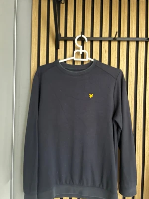 Mörkblå sweatshirt från Lyle & Scott - Snygg mörkblå sweatshirt från Lyle & Scott med klassisk rund hals och gul logotyp på bröstet. Tröjan har långa ärmar och ribbade muddar vid ärmslut och nederkant. Perfekt för dig som gillar stilrena och bekväma plagg. Pris kan diskuteras.
