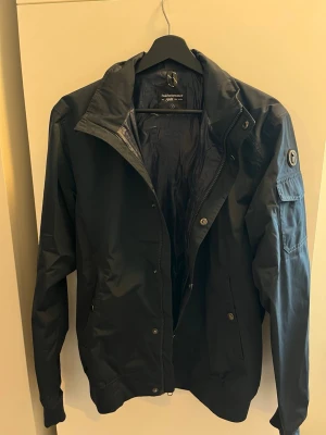 Mörkblå jacka från Peak performance  - Snygg mörkblå jacka från Peak Performance med klassisk passform och diskret logotyp på ärmen. Jackan har dragkedja och knappar framtill, samt fickor med lock på ärmen och sidan. Materialet är lätt och vattenavvisande, perfekt för blåsiga dagar.
