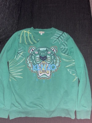 Grön Kenzo sweatshirt med tigertryck - Cool grön sweatshirt från Kenzo med stort broderat tigerhuvud och bladmotiv på framsidan. Tröjan har rund halsringning, ribbade muddar och Kenzo Paris-logga i blått och gult. Perfekt statementplagg för dig som gillar unika prints.