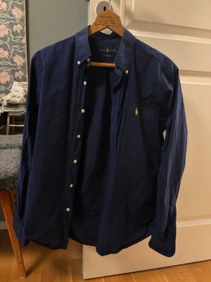 Mörkblå skjorta från Ralph Lauren - Snygg klassisk mörkblå skjorta från Ralph Lauren med button-down krage och en unik gul färg på den ikoniska gula logon broderad på bröstet. Skjortan har långa ärmar, vita knappar och är tillverkad i mjuk bomull. Perfekt för en stilren och avslappnad look.        Nypris 1800kr. Slimfit - M