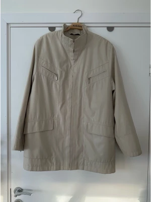 Beige jacka med ståkrage - Concept UK - storlek L - Cool beige jacka från Concept UK med halvglansig finish i storlek L. Jackan har två bröstfickor med dragkedja och två öppna fickor nedtill. Det finns även en innerficka med dragkedja och en ficka för mobilen. Dekorativa dragkedjor vid ärmsluten. Massa fina detaljer både på utsida och insida, kvalitetsjacka. Mycket bra skick, som ny.