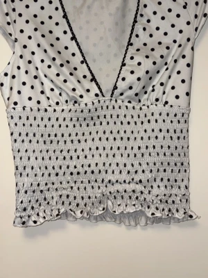 Prickig blus med scrunch och v-ringning  - Snygg vit blus med svarta prickar och djup V-ringning dekorerad med spets. Blusen har smockad midja och volangkant nedtill. Tillverkad i ett glansigt, mjukt material. Fin på men kommer aldorg till användning. Storlek L men kan passa M också pga scrunched i midjan. 