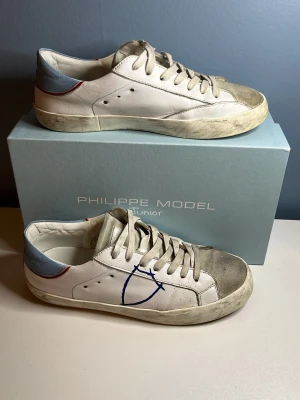 Philippe Model vita sneakers med blå detalj - Snygga vita sneakers från Philippe Model med blå och röda detaljer på hälen samt en blå logga på sidan. Skorna har lågt skaft, beige mockadetaljer på tån och är tillverkade i läder med gummisula. Perfekta för dig som gillar en clean men ändå unik look.