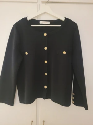 Mörkblå kofta från Busnel med guldknappar - Elegant Marine kofta från Busnel med guldfärgade knappar. Koftan har en rund halsringning och långa ärmar med dekorativa knappar vid ärmsluten. Det finns även två mindre fickor framtill.  Färg: Marine men lutar mer åt svart ? Det är svårt att få den rätta färgnyans när man fotar mörka kläder. Mått: längd 55 cm. Ärmlängd 55 cm.  Koftan var aldrig använd den är helt ny!