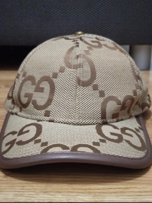 Beige keps med GG-mönster - Säljer en beige keps med det klassiska GG-mönstret i  brunt över hela kepsen. Kepsen har böjd skärm med brun kant, Snygg och tidlös accessoar för dig som gillar exklusiv streetwear.