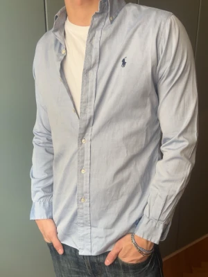 Ljusblå skjorta från Ralph Lauren - Skjorta från Ralph Lauren, inga defekter. Modellen är 187 cm, väger 80 kg och brukar klä i M.   Storlek på skjorta M Mått -|-  Axelmått: 42 cm  Längd: 76 cm  Byst: 54 cm  Armar: 63 cm   Leverans sker dagen efter senast. Skriv om du har nån fråga✨