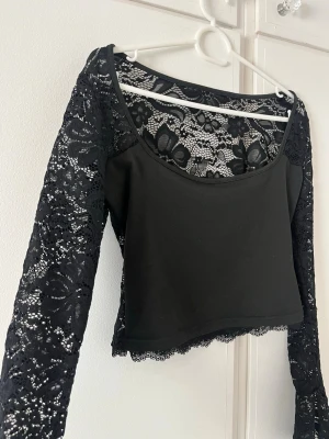 Svart topp i spets - Svart croppad topp med långa ärmar i spets och snygg blomspets över hela ryggen. Urringad framtill och med fina spetsdetaljer längs nederkanten. Perfekt för dig som vill ha en cool och elegant look. 