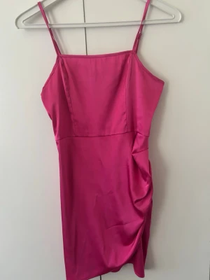 Rosa satin kortklänning - Snygg rosa kortklänning i satin med smala axelband och lätt rynkad detalj på sidan. Klänningen har en enkel, figurnära passform och slät yta. Perfekt för dig som vill sticka ut med en färgstark look.