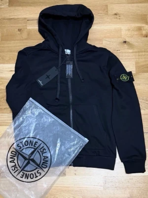 Svart hoodie från Stone Island - Svart hoodie från Stone Island med dragkedja och klassisk kompasspatch på vänster ärm. Tröjan har huva med snören och ribbade muddar. Materialet är mjukt och bekvämt, perfekt för en avslappnad look. Medföljer originalpåse från Stone Island.