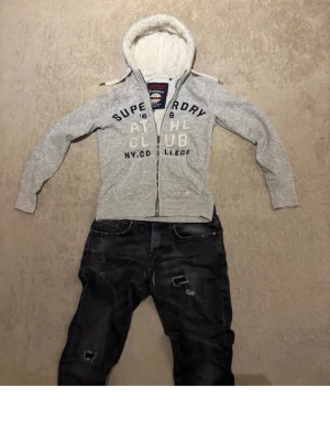 Grå hoodie från Superdry med tryck - Säljer en grå hoodie från Superdry med vitt teddyfoder i huvan och stort tryck på bröstet. Hoodien har dragkedja, två fickor fram och ribbade muddar. Perfekt för dig som gillar sportig och avslappnad stil.