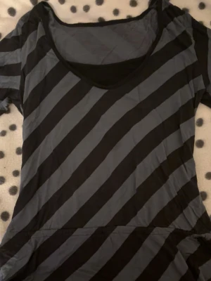 Randig topp med snörning - Säljer en svart och grå randig topp med snörning i sidan. Toppen har korta ärmar och en rundad halsringning. Perfekt för dig som gillar en avslappnad och cool stil.