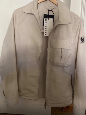 Beige jacka från Belstaff - Snygg beige jacka från Belstaff med klassisk krage och bröstficka med lock. Jackan har dragkedja framtill och ett stilrent märke på ärmen. Materialet är lätt och passar perfekt för lager-på-lager. Perfekt för dig som gillar stilrena och tidlösa plagg. Köper på Johnells. 