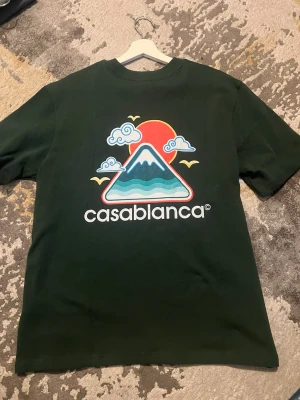 Grön Casablanca t-shirt med tryck - Mörkgrön t-shirt från Casablanca med stort färgglatt tryck framtill som visar en bergsillustration, sol, moln och fåglar. T-shirten har rund hals och korta ärmar. Perfekt för dig som gillar streetwear och grafiska prints. Tröjan är helt ny , inte använd en endaste gång, taggen finns fortfarande kvar. Storlek L. Materialet är ett tjockt och skönt kvalitets plagg.  Pris kan diskuteras!