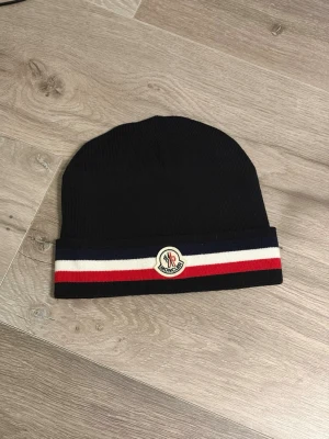Svart Moncler mössa med ränder - Cool svart mössa från Moncler med ribbstickad struktur. Mössan har en bred kant med blå, vit och röd rand samt Moncler-logga framtill. Perfekt för dig som vill ha en stilren accessoar med exklusiv känsla. Materialet är troligtvis bomull eller ull.