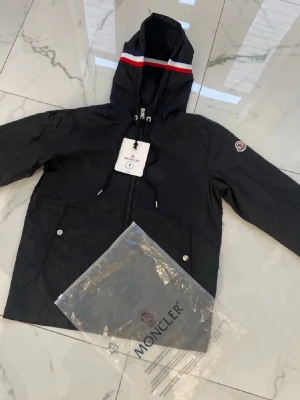 Svart Moncler vindjacka med huva - Svart vindjacka från Moncler med huva och dragkedja. Jackan har vita och röda detaljer på huvan samt Moncler-logga på ärmen. Materialet är lätt och vattenavvisande, perfekt för blåsiga dagar. Två fickor med knappar framtill och justerbar huva.
