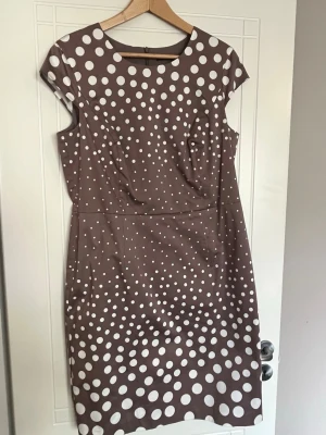 Brunprickig klänning från Esprit - Snygg brun klänning från Esprit med vita prickar i olika storlekar. Modellen är knälång med korta ärmar och rund halsringning. Perfekt för dig som gillar retrovibbar och ett lekfullt mönster. Materialet känns mjukt och följsamt.