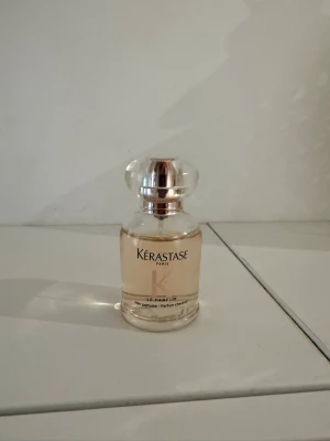 Kérastase Le Parfum Eau de Parfum - Elegant parfymflaska från Kérastase med transparent glas och rund, silverfärgad kork. Innehållet är ljusrosa och flaskan har en klassisk, lyxig design som passar perfekt på sminkbordet. Märket är känt för sina exklusiva dofter och stilrena förpackningar.