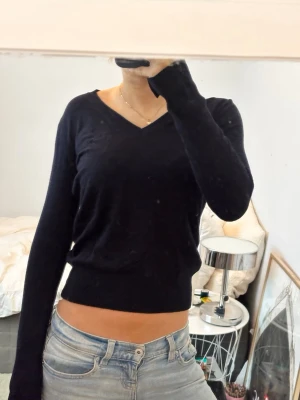 Mörkblå V-ringad stickad tröja - Snygg svart stickad tröja med V-ringning och långa ärmar. Tröjan har en smal passform och kan bäras både på axeln eller off-shoulder för en mer avslappnad look. Perfekt att matcha med jeans för en enkel men stilren outfit.