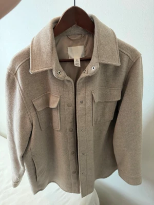 Beige overshirt med fickor - Stilren beige overshirt i mjukt material med två stora bröstfickor och tryckknappar framtill. Klassisk krage och raka ärmar ger en clean look. Perfekt lager-på-lager-plagg för dig som gillar minimalistisk stil.