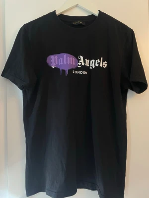 Svart Palm Angels t-shirt London - Svart t-shirt från Palm Angels med tryck i vitt och lila på bröstet där det står 'Palm Angels London'. Klassisk passform och rund hals. Tillverkad i 100% bomull för en skön känsla. Perfekt för dig som gillar streetwear och vill sticka ut. (Inte äkta) men går att skanna QR koden. Det är XLen tycker den sitter som en L eller M. 