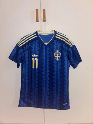 Blå Sverige fotbollströja Adidas - Säljer en blå fotbollströja från Adidas med Sveriges landslagsmärke och nummer 11 på bröstet och ryggen. Tröjan har Elanga tryckt på ryggen, gula detaljer och mönster längs hela tyget. Perfekt för dig som vill representera Sverige i VM. Säljer då den satt lite tajt på mig.