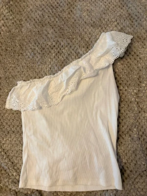 Vit oneshoulder topp med volang - Snygg vit oneshoulder topp med bred volangkant i broderad spets. Toppen är ribbad och har en axel bar, vilket ger en trendig och feminin look. Perfekt för varma dagar eller sommarens alla tillfällen.