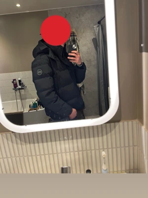 Svart dunjacka från Canada Goose - Säljer en svart dunjacka från Canada Goose med huva och klassisk patch på ärmen. Jackan är tjock, har dragkedja och knappar framtill samt flera fickor. Perfekt för kalla vinterdagar och riktigt snygg streetstil.