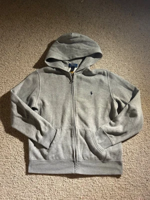 Grå zip hoodie från Polo Ralph Lauren - Klassisk grå hoodie med dragkedja från Polo Ralph Lauren. Tröjan har huva, två fickor fram och den ikoniska lilla broderade loggan på bröstet. Tillverkad i mjuk bomullsblandning och har ribbade muddar vid ärmslut och nederkant. Storlek 14-16 år=L barnstorlek.