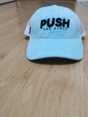 Ljusblå keps med PUSH THE LIMIT - Cool ljusblå keps med svart broderad text 'PUSH THE LIMIT' framtill och bak. Kepsen har vit mesh på sidorna och justerbar snapback. Perfekt för dig som gillar sportig och avslappnad stil. Har inte låda eller kvitto