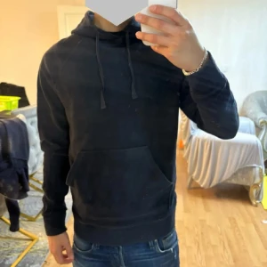Mörkblå hoodie med ficka framtill - Säljer en mörkblå hoodie med klassisk känguruficka och justerbar huva med snören. Hoodien har ribbade muddar vid ärmslut och nederkant. Perfekt för en avslappnad och stilren look.