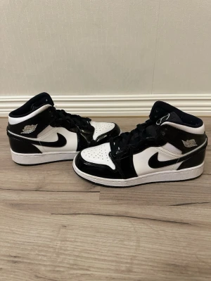 Nike Air Jordan 1 Mid svart/vit - Säljer ett par Nike Air Jordan 1 Mid sneakers i svart och vitt med klassisk Swoosh-logga på sidan och Air Jordan Wings på ankeln. Skorna har en kombination av läder och syntetmaterial, perforerad tå och svarta skosnören. Perfekt för dig som gillar ikoniska sneakers.