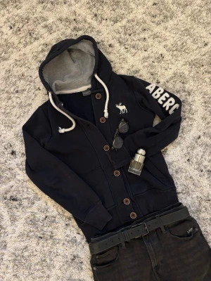 Mörkblå hoodie från Abercrombie & Fitch - Cool mörkblå hoodie med knappar och vit broderad älg på bröstet. Stor vit logga på ärmen och grått innerfoder i huvan. Tjocka vita snören och ribbade muddar | Size S passar upp till 175! | Inga defekter!