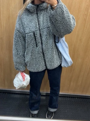 Grå teddyjacka med dragkedja - Mysig grå teddyjacka i oversized modell med hög krage och hel dragkedja framtill. Jackan har en bröstficka med dragkedja och två sidofickor. Perfekt för kalla dagar och ger en cool, avslappnad look.